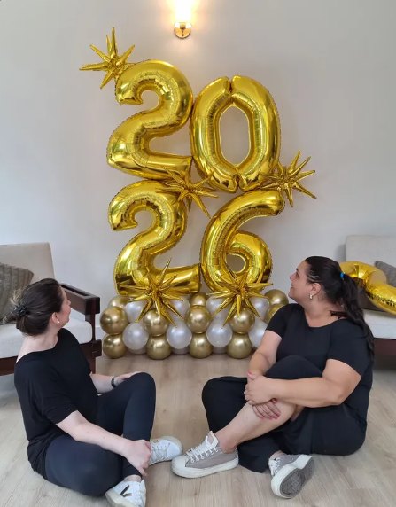 As duas sócias da Alliance Balões sentadas no chão diante de balões dourados que formam 2026